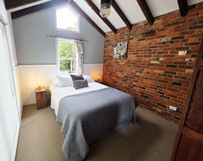 A bedroom with a bed, a nightstand, a window with curtains, a brick wall, and a ceiling with exposed beams. - Cascade Cottage | Circa 1840 (Hobart City)