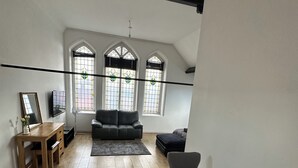 Ein geräumiges Wohnzimmer mit Holzboden, einem grauen Sofa, einem runden Tisch mit einer Vase und einem Holzbüro mit Spiegel und Vase.