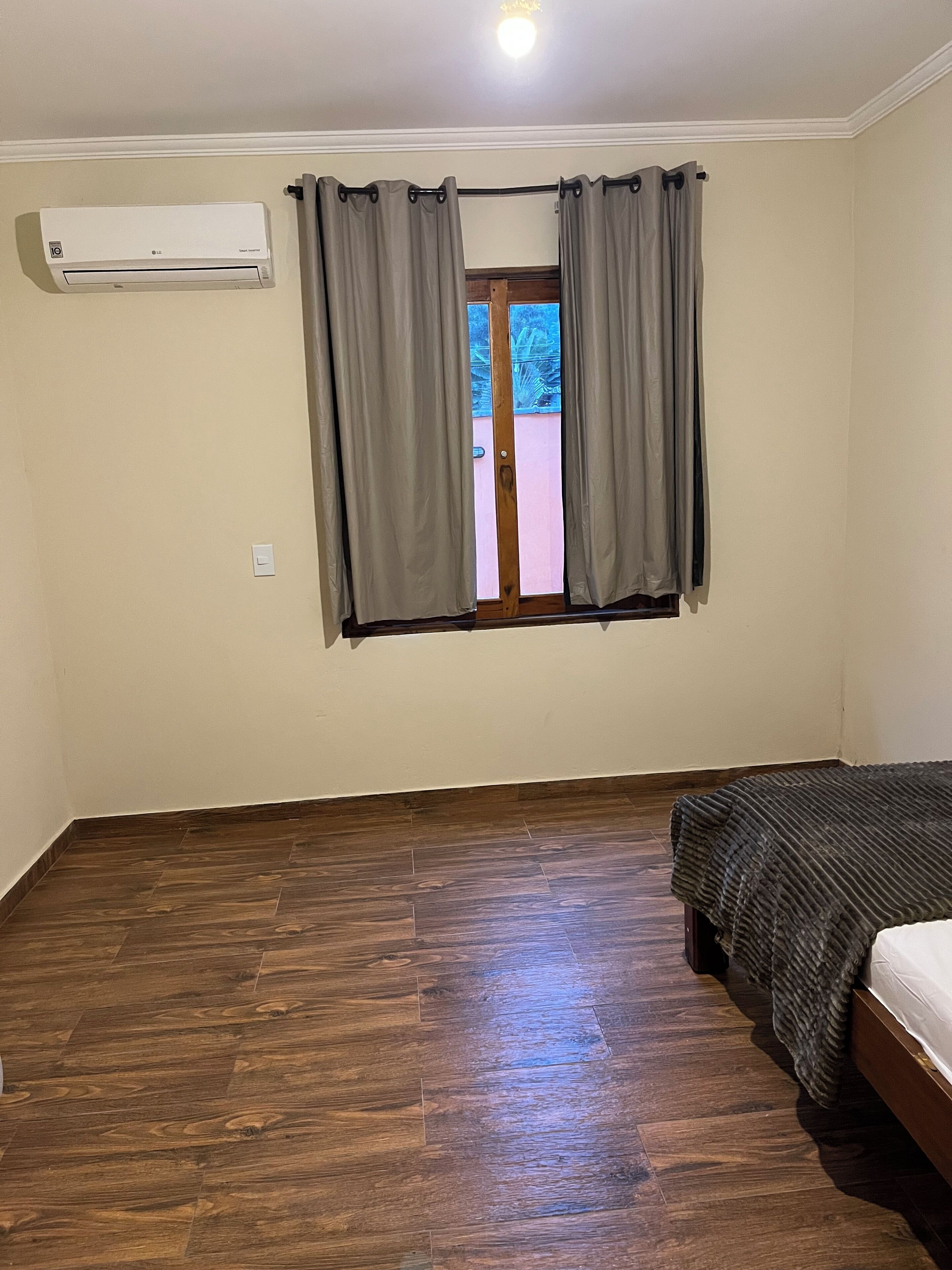 Una habitación con piso de madera, una cama con cubrecama oscuro, aire acondicionado y una ventana con cortinas grises.