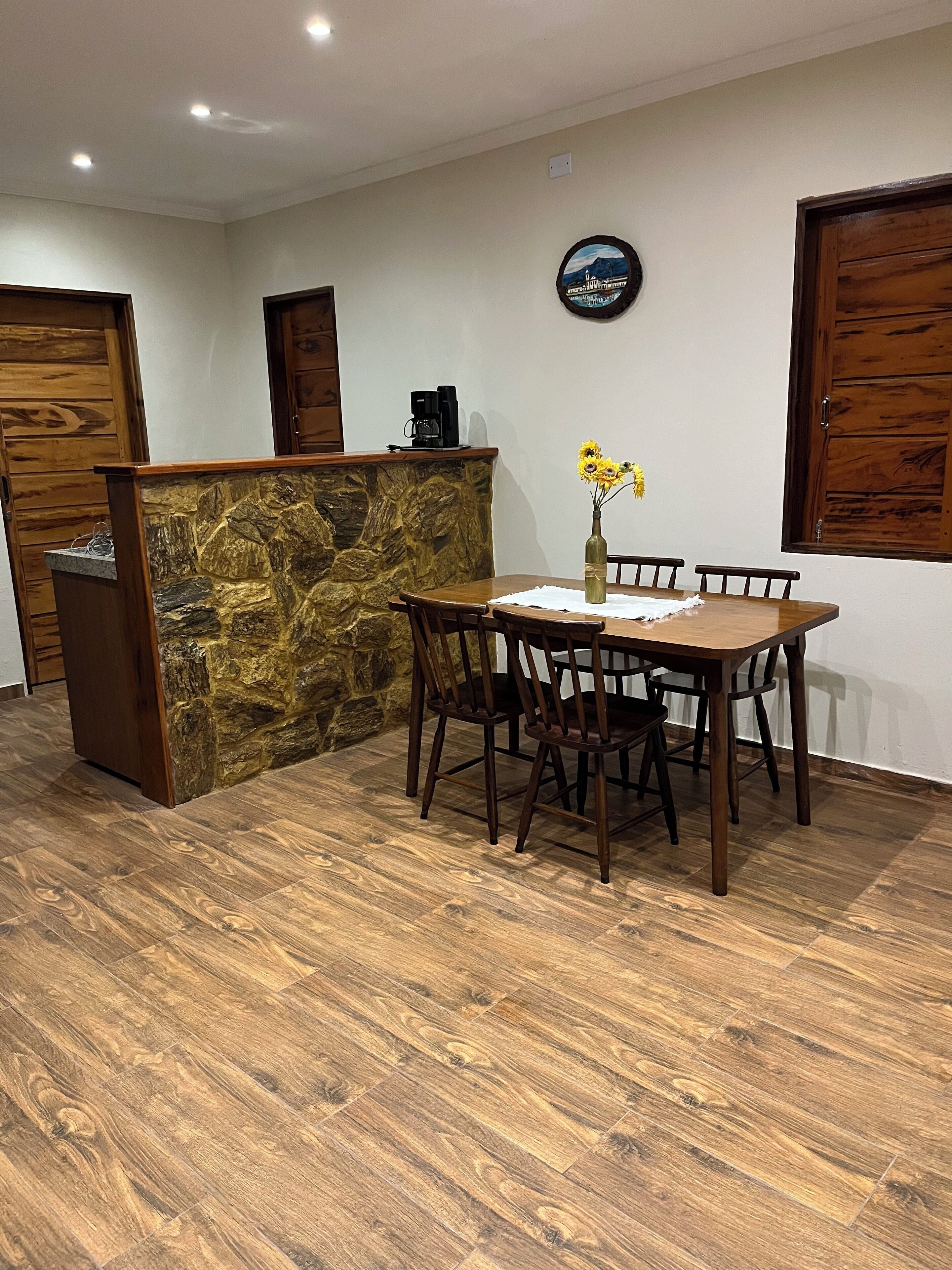 A dining area with a stone accent wall, wooden table and chairs, a coffee machine, and a vase with flowers.