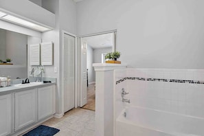 Une salle de bain moderne avec un lavabo blanc, un miroir et une baignoire. Une porte donne accès à une autre pièce, et une petite plante est posée sur le comptoir.