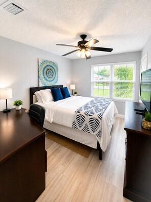 A bedroom with a bed, a ceiling fan, a window with blinds, a wall art piece, and a desk with a lamp.