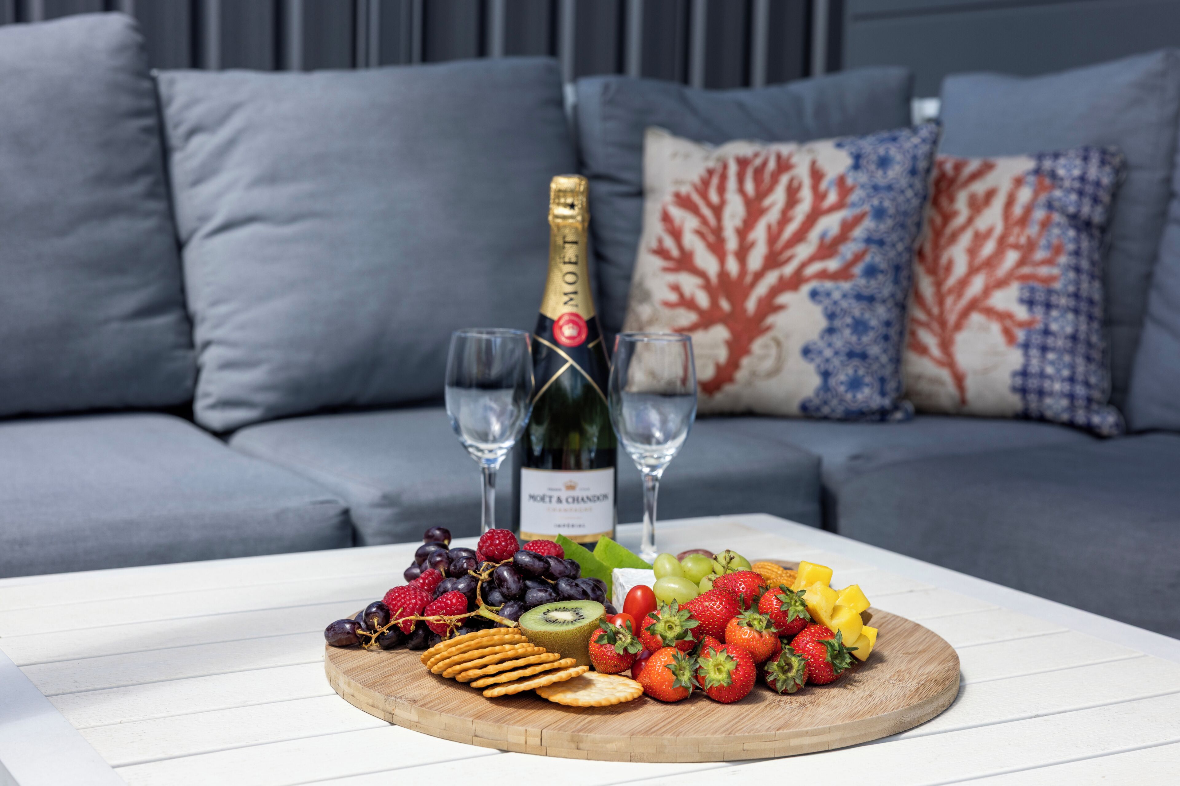 A wooden board with fruits and crackers on a white table, a bottle of champagne, and two glasses in front of a grey sofa with patterned cushions.