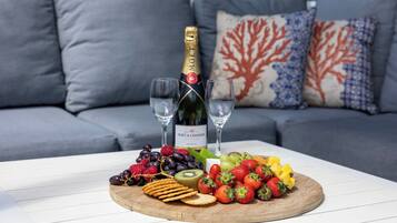 A wooden board with fruits and crackers on a white table, a bottle of champagne, and two glasses in front of a grey sofa with patterned cushions.