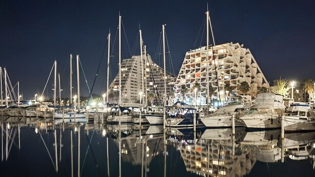Uma marina à noite com vários barcos atracados, um edifício grande com design arquitetônico único e uma superfície de água calma refletindo a paisagem.