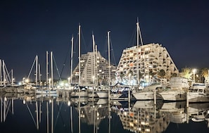 A marina at night with multiple boats docked, a large building with a unique architectural design, and a calm water surface reflecting the scene. - 1 bedroom amazing apartment in La Grande-Motte (La Grande-Motte)