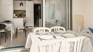 Uma sala de jantar com uma mesa posta para quatro pessoas, cadeiras brancas e uma cozinha ao fundo.