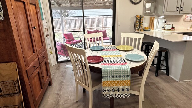 A dining area with a table set for four, surrounded by white chairs, a patterned tablecloth, and a view through a glass door to an outdoor patio with chairs and a fence. - Kechi Kottage- Sept. special- 20% off 2+ ni stay! Ask host. Pet friendly. Safe! (Kechi)