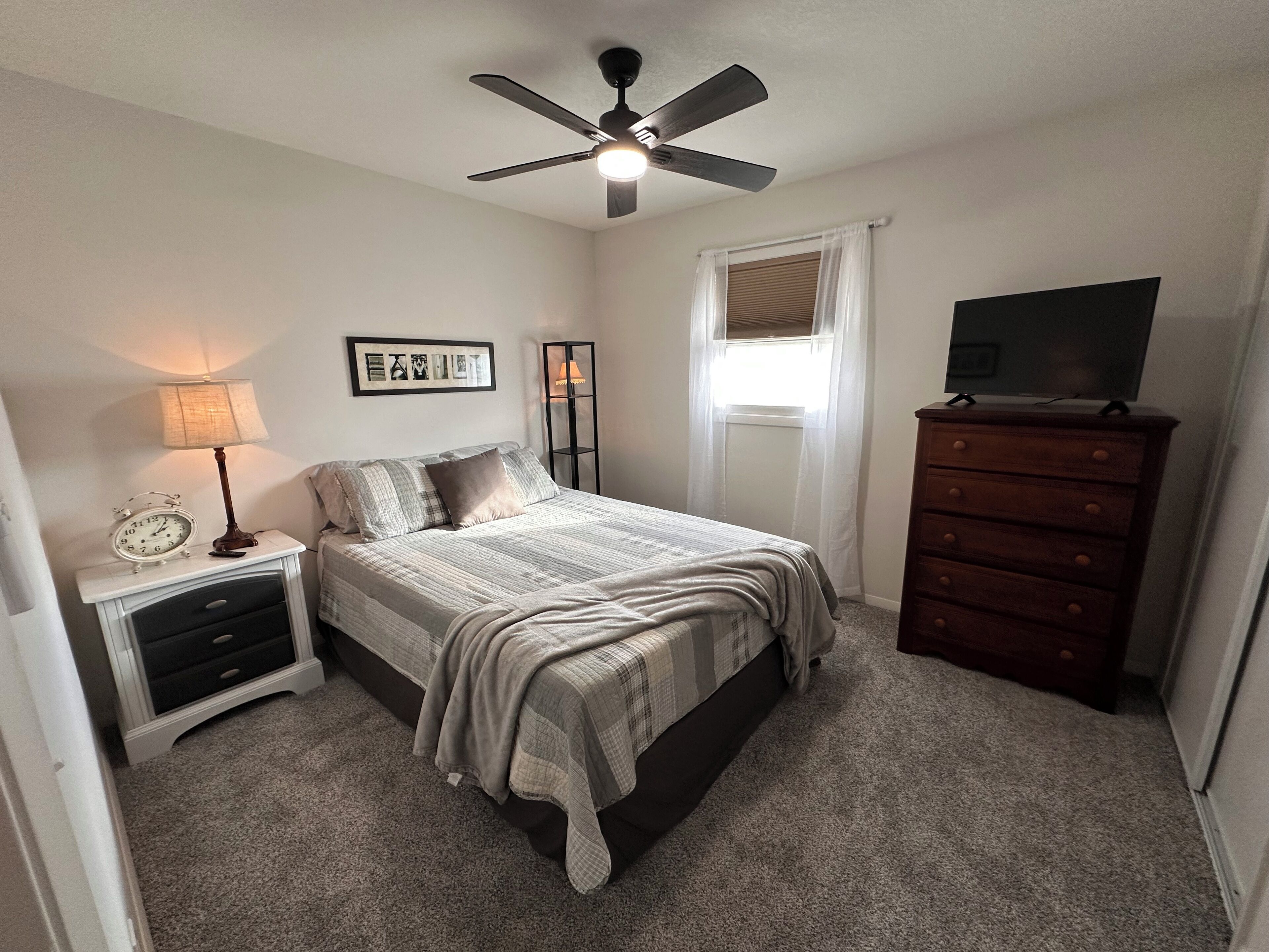 Une chambre à coucher comprenant un lit, une commode, une table de chevet, une lampe, un ventilateur de plafond, une fenêtre avec des rideaux et un tableau encadré accroché au mur.