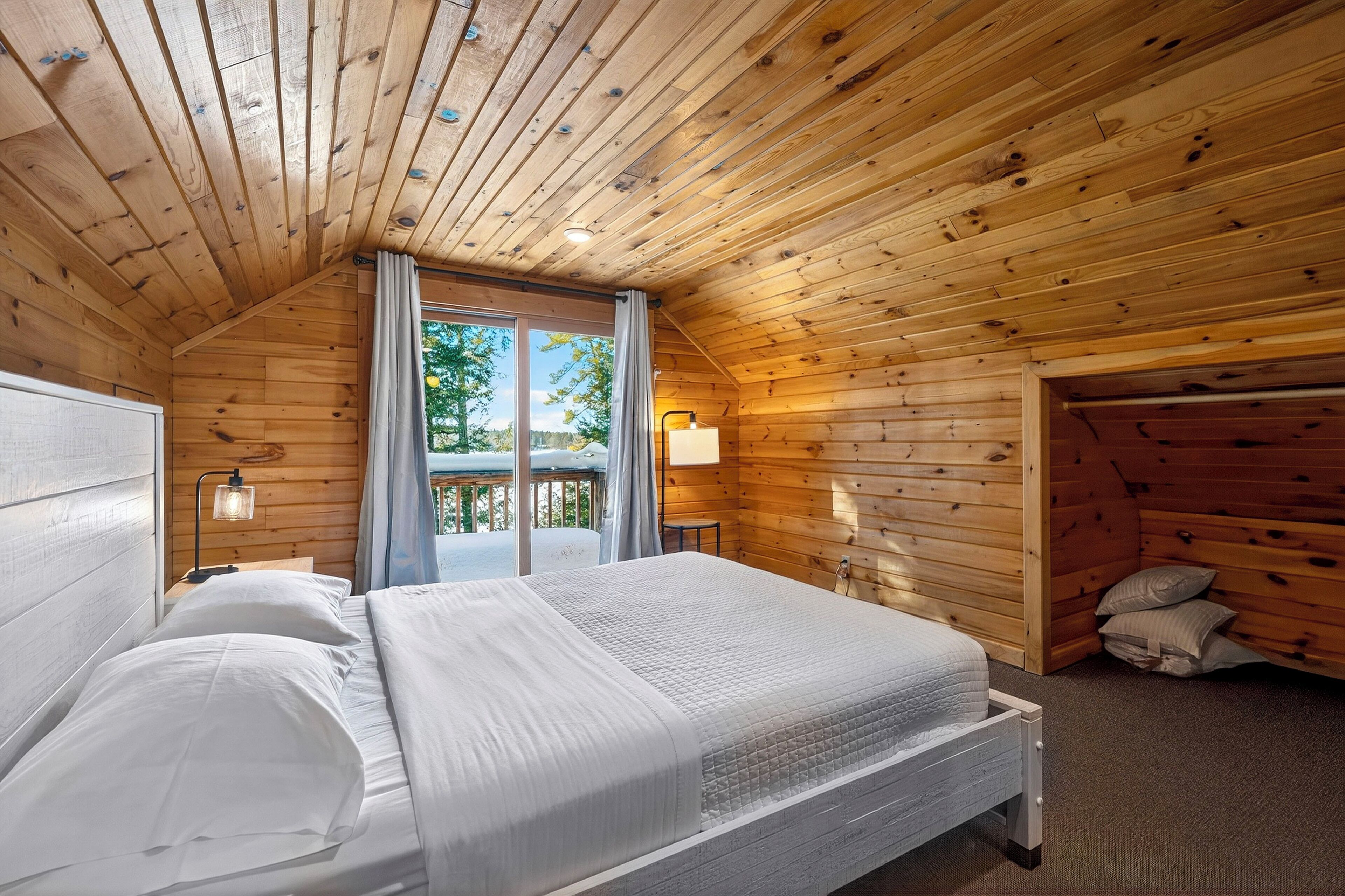 Une chambre à coucher dans une cabane en bois, avec un lit, deux lampes de chevet, une fenêtre avec des rideaux et une vue sur l’extérieur.