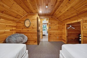 Un dormitorio tipo cabaña de madera con dos camas, un almohadón mullido, una puerta de madera y un pasillo que conduce a un baño.