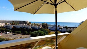 Un balcón con una sombrilla amarilla, sillas blancas y vistas al mar y edificios.