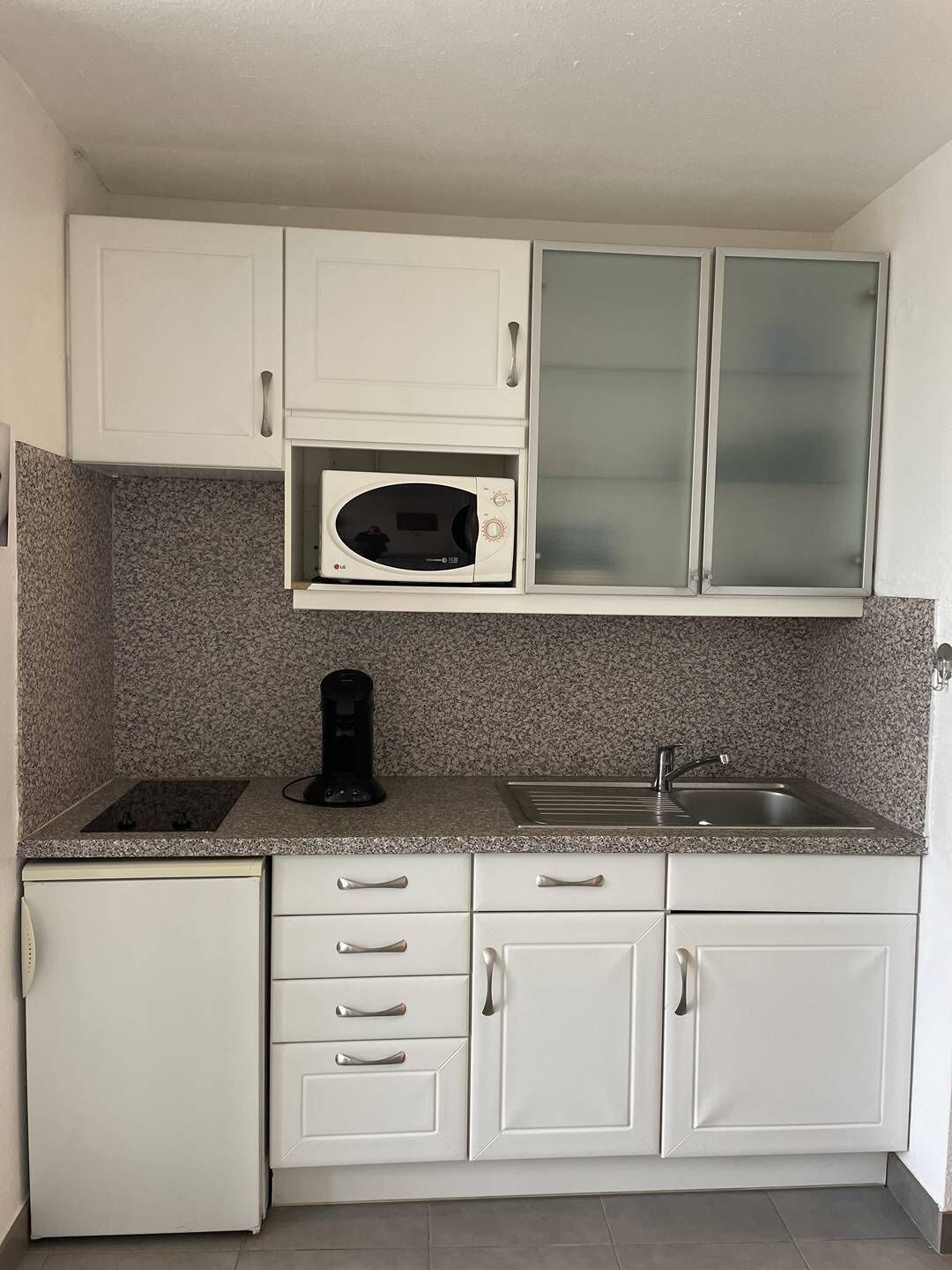 A compact kitchen with white cabinets, a microwave, a coffee maker, a sink, and a small refrigerator.