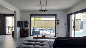 Een moderne woonkamer met een bank, een salontafel en een televisie. Er is een schuifdeur die leidt naar een terras buiten met een tafel en stoelen.