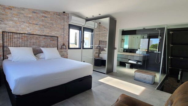 Een moderne slaapkamer met een groot bed, een bakstenen muur en een zichtbare badkamer door een glazen deur.