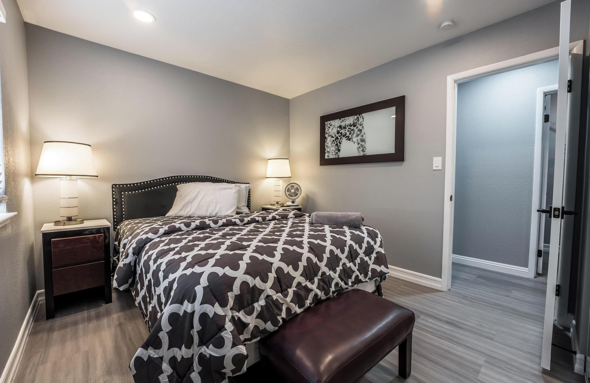 A bedroom with a bed, two bedside tables with lamps, a bench, a door, and a wall-mounted artwork.