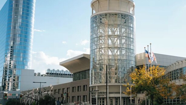 Un bâtiment moderne doté d’une tour cylindrique en verre, d’une structure à toit plat et d’un mât sur lequel flottent des drapeaux.