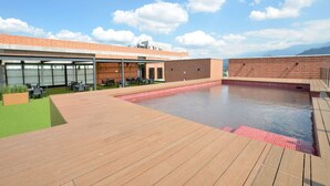 Ein Poolbereich auf dem Dach mit Holzterrasse, Backsteinmauer und einer überdachten Lounge mit Sitzgelegenheiten im Freien.