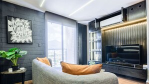 Uma sala de estar moderna com um sofá cinza, uma TV de tela plana e uma janela grande com vista para a cidade.