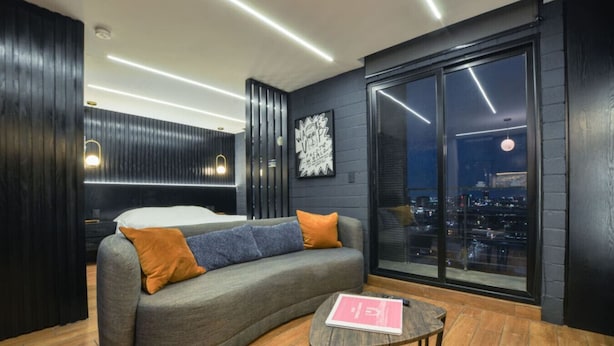 Sala de estar moderna com sofá cinza curvo, uma mesa de madeira pequena e vista para a cidade através de janelas amplas.