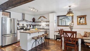A modern kitchen with a central island, wooden bar stools, a dining table with chairs, and a hanging light fixture.