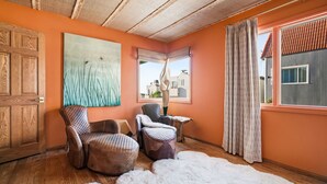Ein Zimmer mit Holzboden, orangefarbenen Wänden, einem großen Fenster mit Vorhängen, einer Holztür und zwei Ledersesseln.