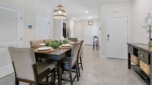 A modern dining area with a long table set for six, surrounded by wicker chairs, and a hanging light fixture above. - Palm Villa: 5 beds Playground, UNF, Mayo, Downtown (Jacksonville)