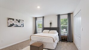 A bedroom with a bed, a small ottoman, a nightstand, a lamp, a window with curtains, a wall-mounted plant, and a painting. - Palm Villa: 5 beds Playground, UNF, Mayo, Downtown (Jacksonville)