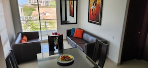 A modern living room with a glass coffee table, two dark sofas with colorful cushions, a framed painting, and a view of buildings through large windows. - Palmagua (Barranquilla)