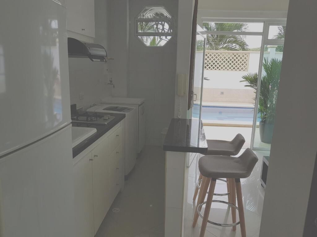 Une cuisine étroite avec des meubles blancs et un plan de travail noir, un petit coin bar avec des tabourets, et une vue sur une piscine à travers une porte vitrée.
