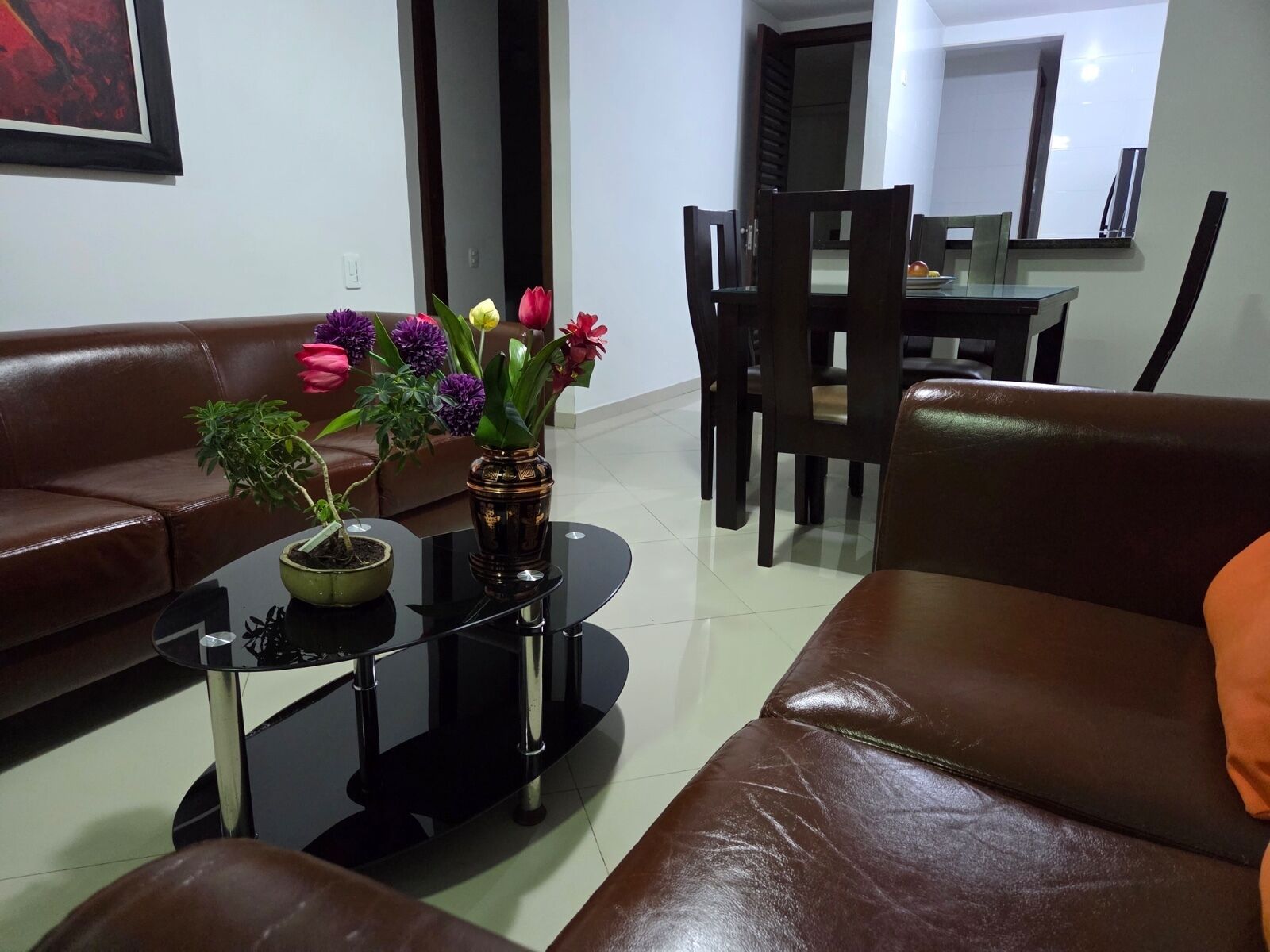 Un salon avec un canapé en cuir marron, une table basse en verre ornée d’une plante en pot, un coin repas avec une table et des chaises en bois, et un tableau encadré accroché au mur.
