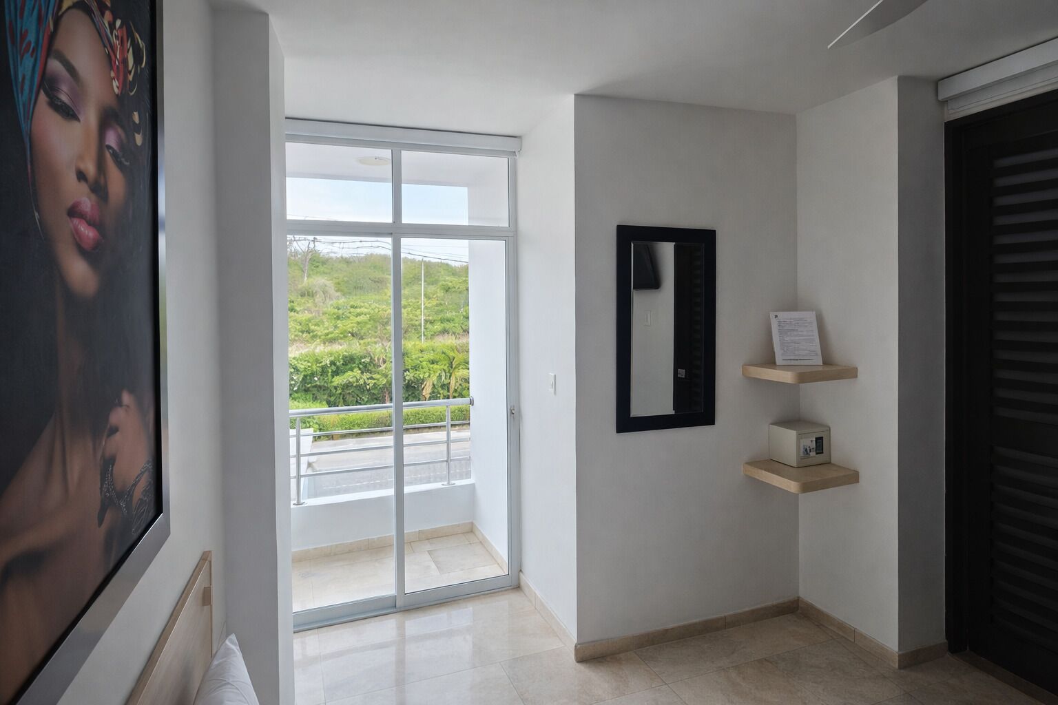 Un interior moderno con una puerta corrediza de vidrio que da a un balcón, un espejo enmarcado y un pequeño estante con un dispositivo.