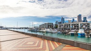Un puerto deportivo con barcos amarrados, un horizonte urbano al fondo y un paseo pavimentado en primer plano.