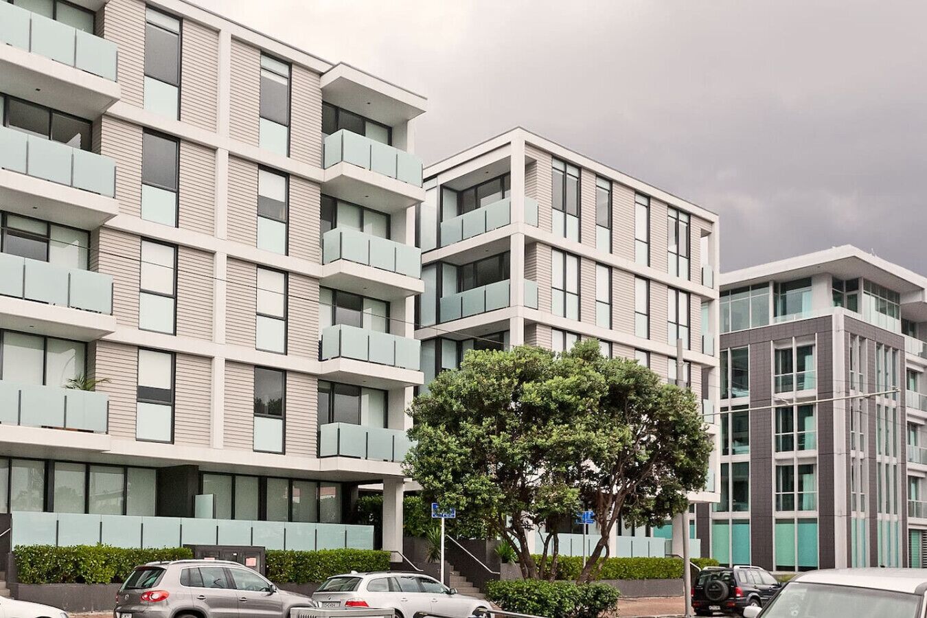 A multi-story residential building with balconies, a tree in front, and parked cars.
