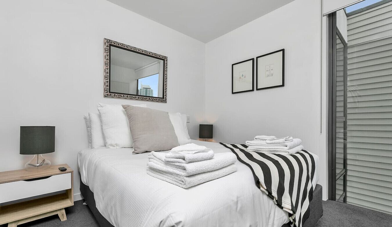 A bedroom with a bed, a nightstand, a lamp, a mirror, and a window with blinds.