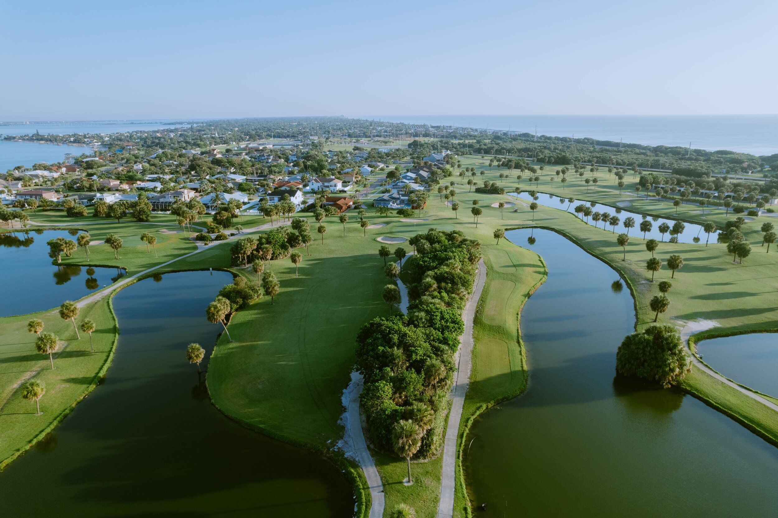 Un campo de golf con obstáculos acuáticos, un sendero para pasear y una zona residencial al fondo.