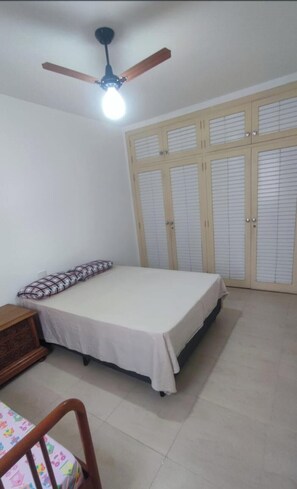 Ein Schlafzimmer mit Bett, Nachttisch, Holzstuhl, Deckenventilator und Kleiderschrank mit Klappläden.