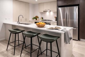 A modern kitchen with a central island, bar stools, a stainless steel refrigerator, and a bowl of oranges. - Brisbane CBD 2BR Gem w/ Skyline Views + Parking (Brisbane)