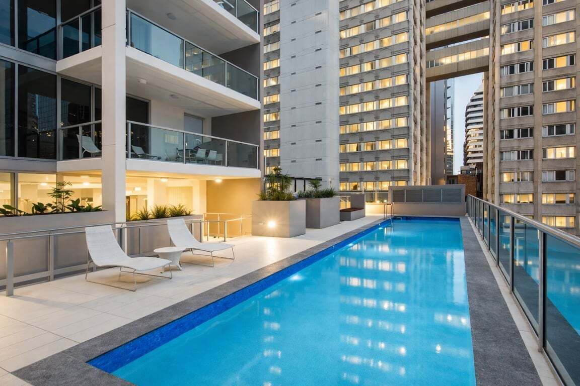 A rooftop pool with lounge chairs, surrounded by modern buildings with balconies and large windows.