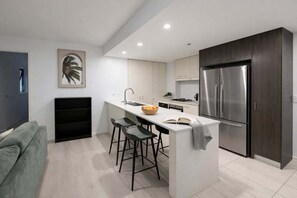A modern kitchen with a central island, stainless steel appliances, and a grey sofa in the adjacent living area. - Brisbane CBD 2BR Gem w/ Skyline Views + Parking (Brisbane)