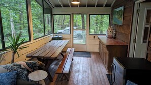 A cabin with a wooden interior, large windows, a dining area with benches, and a view of the forest.