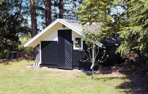 A small, black wooden cabin with a white door and window, surrounded by trees and grass.