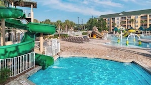 Una piscina con tobogán de agua, una pequeña cabaña y un edificio con balcones al fondo.
