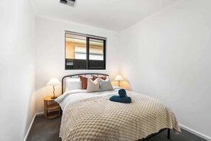 A neatly arranged bedroom with a bed, pillows, a bedside lamp, and a window with blinds. - Stylish 3BR Stay Close to Tea Tree Plaza (Modbury)
