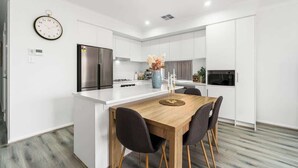 A modern kitchen with white cabinetry, a central island, a stainless steel refrigerator, and a dining area with a wooden table and chairs. - Stylish 3BR Stay Close to Tea Tree Plaza (Modbury)