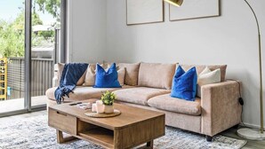A living room with a beige sofa, blue pillows, a wooden coffee table, and a floor lamp. - Stylish 3BR Stay Close to Tea Tree Plaza (Modbury)
