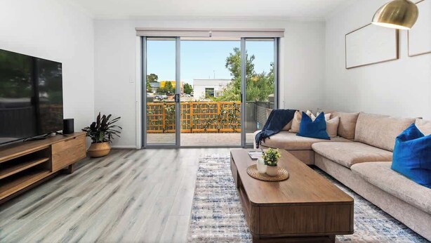 A modern living room with a large TV, sofa, coffee table, and a view through a sliding glass door to an outdoor area. - Stylish 3BR Stay Close to Tea Tree Plaza (Modbury)