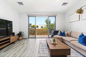 A modern living room with a large TV, sofa, coffee table, and a view through a sliding glass door to an outdoor area. - Stylish 3BR Stay Close to Tea Tree Plaza (Modbury)