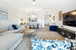 Uma sala de estar moderna com um sofá, um banquinho e uma mesa de centro. Há uma área de jantar com um candelabro e uma cozinha ao fundo.