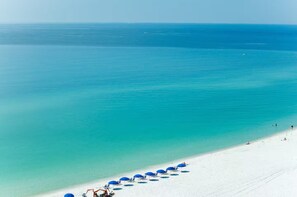 Ein Strand mit weißem Sand und klarem türkisblauem Wasser. Entlang der Küste stehen blaue Sonnenschirme und Liegestühle.